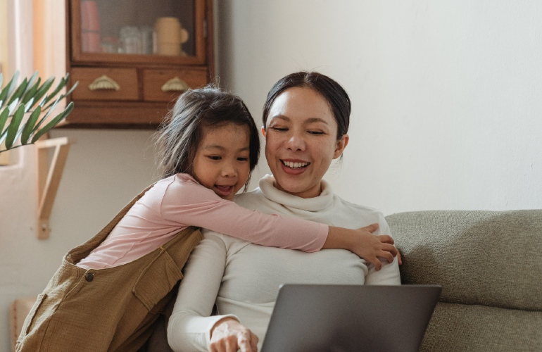 A woman with a laptop on her sofa, while being hugged by a young female child