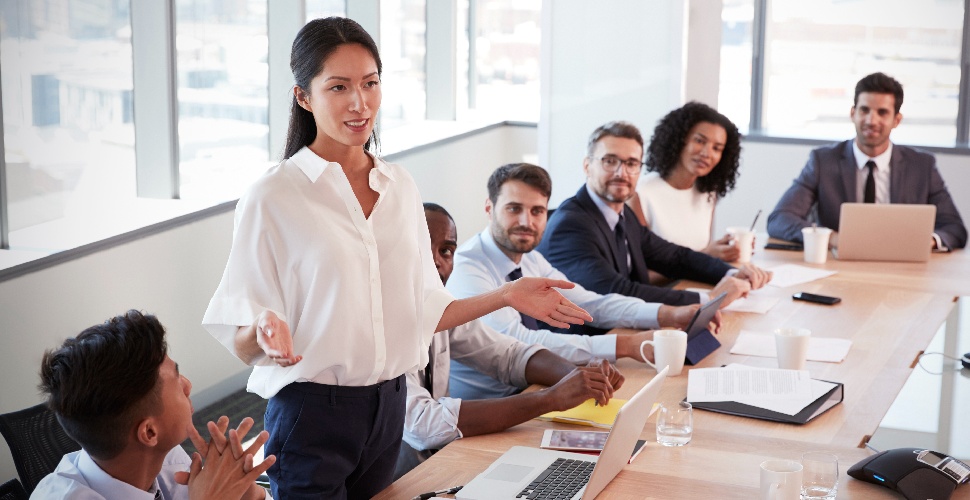 A group of white-collar professional sitting around a desk with one Asian female standing and speaking