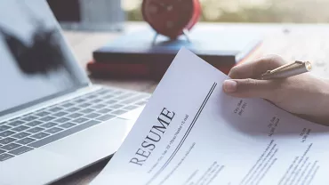 Person holding a resume next to a laptop on a wooden desk.