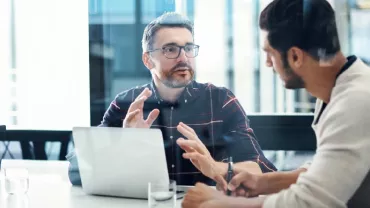 Two people have a discussion in an office with a laptop and notepad.