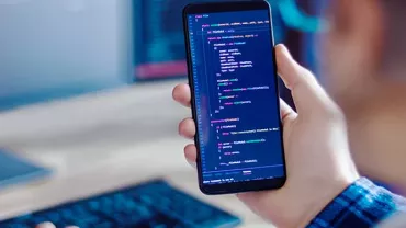 Person holding a phone displaying code, with a blurred computer screen in the background.