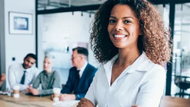 A female professional smiling, on a office background with three people dressed in corporate wear seated behind her