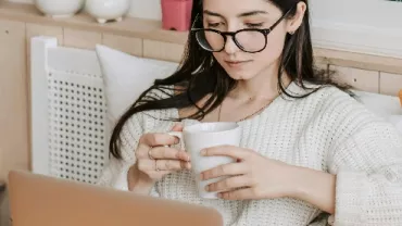 Person sitting on a cozy couch, holding a coffee mug while using a laptop.