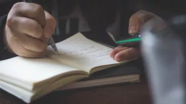 Person writing notes in a journal while holding a smartphone.