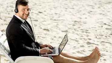 Person in a suit working on a laptop at the beach, wearing headphones.