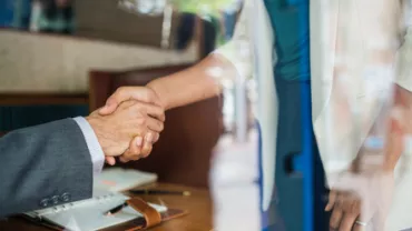 Two people shaking hands over a wooden table, seen through a window.