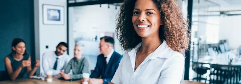 A female professional smiling, on a office background with three people dressed in corporate wear seated behind her